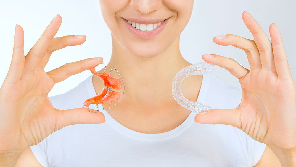 Lady smiling and holding up a wired retainer in one hand and a clear retainer in the other